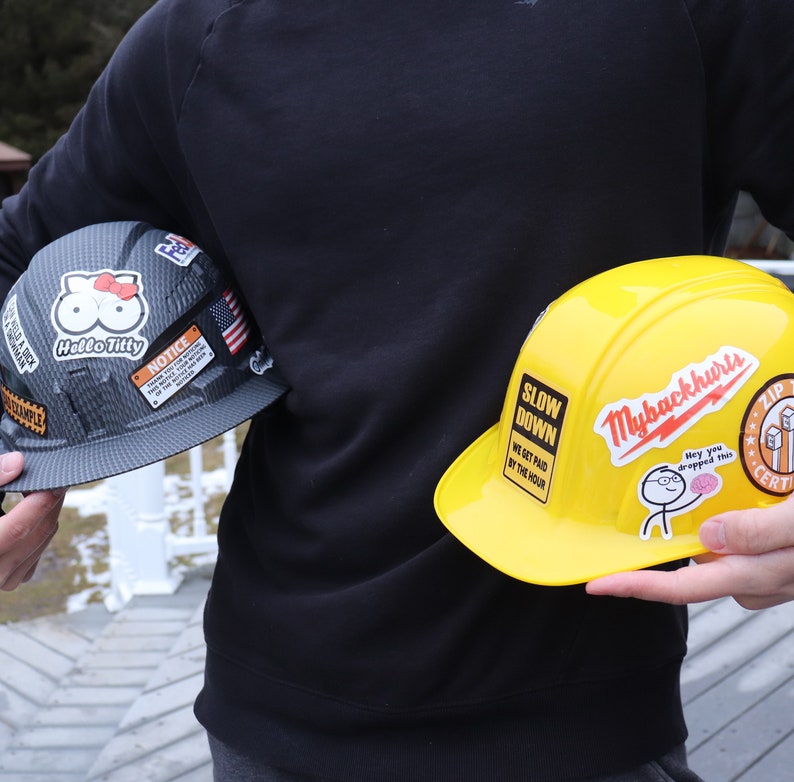 May include: Two hard hats with various stickers. The first hat is black with a Hello Titty sticker and a notice sticker. The second hat is yellow with stickers that say Slow Down, We Get Paid By The Hour, Mybackhurts, Hey You Dropped This, Zip Certified.