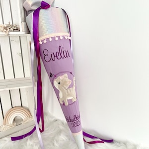 Könnte beinhalten: Eine lavendelfarbene Schultüte mit einem Katzendesign und dem Namen "Evelin". Die Tüte hat einen Regenbogenfarbenen-Deckel, lila Bänder und den Text "Schulkind 2025". Sie ist mit weißem Besatz und Bommeln verziert.