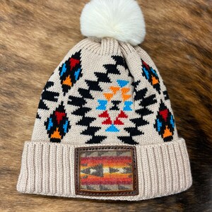 May include: A beige knitted beanie with a white pom pom and a geometric pattern in black, red, orange, and blue. The beanie has a brown leather patch with a colorful woven design.