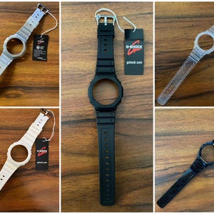 May include: Collection of G-Shock watch bands in various colors, including silver, black, clear, white, and navy blue. Each band has a matching watch face holder and a tag with the G-Shock logo and website.