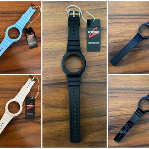 May include: A collection of G-Shock watch straps and cases in various colors, including light blue, white, and black. Each strap has a buckle and a tag with the G-Shock logo and website address. The watch cases have a circular opening for the watch face.