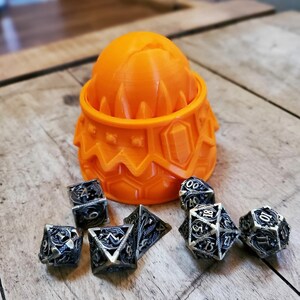 May include: Orange plastic dragon egg container with six metal dice with black and gold designs. The dice are shaped like a pyramid with four sides.