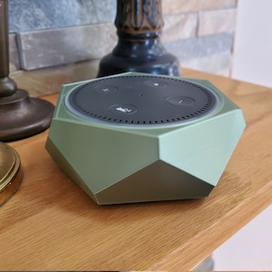 May include: A green hexagonal speaker with a black top and four buttons. The buttons are labeled with icons for volume up, volume down, microphone mute, and a plus sign.