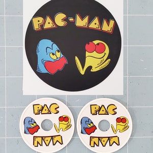 May include: Two white Pac-Man game tokens with a black and yellow Pac-Man character and a blue ghost character. The tokens have a hole in the center. A black and white sticker with the text "PAC-MAN" and a yellow Pac-Man character and a blue ghost character.