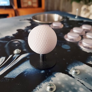 May include: A white, textured, round joystick knob with a black base. The knob is mounted on a black surface with a blue and white abstract design.