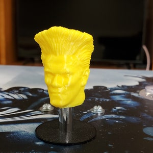 May include: A yellow 3D printed head with a spiky hairstyle, mounted on a black base with a silver metal shaft.