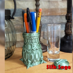 May include: A green, 3D printed, hexagonal patterned pen holder with a lid. The holder is shaped like a vase and has a decorative base. The lid is also hexagonal and has a small handle. The holder is filled with pens and pencils.