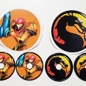 May include: Five round stickers, two featuring a black silhouette of a dragon with a yellow and orange background, and three featuring a cartoon character in a gold and red suit with a blue gun, on a white background.