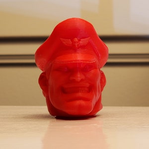 May include: A red 3D printed figurine of a man's head wearing a red cap with a winged emblem. The man has a stern expression with a wide smile and a prominent chin.
