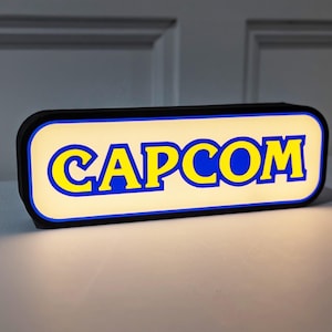 May include: A black rectangular sign with a white background and blue outline. The word "CAPCOM" is written in yellow letters.