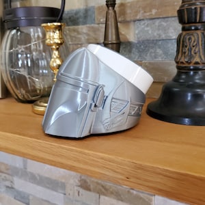 May include: A silver Mandalorian helmet with a white base, featuring a raised design of the Mandalorian symbol. The helmet is sitting on a wooden surface.