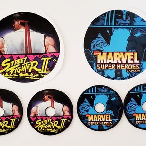 May include: Set of six round arcade game control panel overlays. Two feature a black and white image of Ryu from Street Fighter II. Two feature a blue, black, and white image of Wolverine and Sabretooth from Marvel Super Heroes. Two feature a blue, black, and white image of Thanos and Wolverine from Marvel Super Heroes.