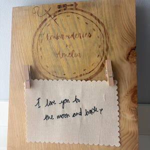 May include: A rectangular wooden board featuring a wood-burned embroidery hoop design with the text Embroidries by Amelia. A rectangular piece of off-white fabric with scalloped edges is attached with two wooden clips, displaying the embroidered text I love you to the moon and back.
