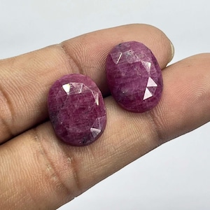 Puede incluir: Dos gemas ovaladas, de color rojo intenso, con múltiples facetas. Las gemas tienen una superficie pulida que refleja la luz. Las piedras se sostienen en una mano, mostrando su color y talla. Estas gemas son adecuadas para la fabricación de joyas.