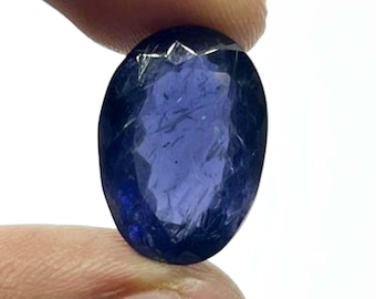 May include: A faceted, oval-shaped, deep blue gemstone. The gem is held between fingers against a white background. The facets create a play of light and shadow, enhancing the stone's brilliance. The overall appearance suggests a precious stone, possibly for jewelry.