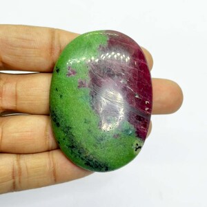 May include: Oval-shaped gemstone with a polished surface. The stone features a vibrant green hue on one side, transitioning to a deep red-purple on the other. The gemstone is held in a hand, showcasing its size and shape.