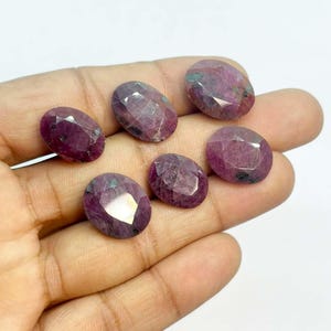 Peut inclure: Sept pierres précieuses rubis ovales et facettées, d'un rouge-violet profond avec des inclusions internes. Les surfaces polies reflètent la lumière. Les pierres sont présentées sur une main, mettant en valeur leur aspect et leurs variations naturelles.