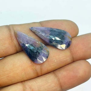 May include: Two faceted, teardrop-shaped gemstones. The stones have a lavender hue with darker blue and black inclusions. The gems are held in a hand, showcasing their polished surfaces and unique color variations. These stones are likely for jewelry making.