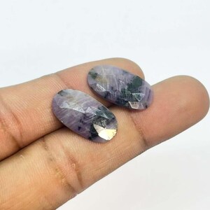 Könnte beinhalten: Zwei ovale, facettierte Edelsteine mit violetten und schwarzen Farbtönen. Die Edelsteine haben eine polierte, reflektierende Oberfläche und werden vor einem neutralen Hintergrund präsentiert. Diese Edelsteine werden wahrscheinlich für die Schmuckherstellung verwendet.