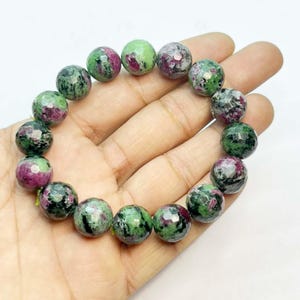 Puede incluir: Un brazalete de cuentas redondas y facetadas. Las cuentas son verdes y rojo oscuro, con un aspecto moteado. El brazalete se sostiene en una mano sobre un fondo blanco. Es una pieza de joyería.