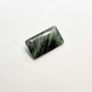 May include: A rectangular, faceted gemstone with a mix of green and black hues. The stone has a polished, reflective surface and a geometric shape. The gemstone is set against a plain white background.
