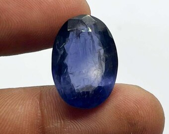 May include: A faceted, oval-shaped, deep blue gemstone. The gem is held between two fingers, showcasing its polished surface and internal clarity. The stone's color ranges from a dark, saturated blue to lighter shades.