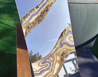 May include: Rectangular mirror with a decorative design. The mirror features a blue sky reflection and is adorned with gold, white, and purple resin, creating a geode-like pattern. The edges are embellished with gold-colored accents.