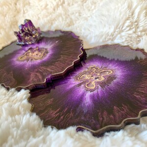 May include: Two purple and gold resin coasters with a glittery finish. The coasters have a unique agate design and are perfect for adding a touch of elegance to any home.