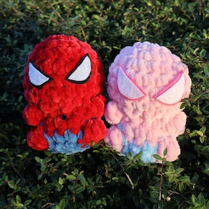 May include: Two plush toys, one red and one pink, resembling superhero characters. The red toy has white eye accents and blue accents. The pink toy has pink eye accents and blue accents. Both are set against a backdrop of green foliage.