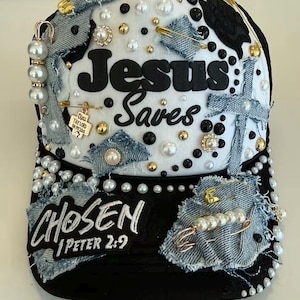 Puede incluir: Una gorra de béisbol negra y blanca con el texto Jesus Saves y Chosen 1 Peter 2:9. La gorra está decorada con parches de tela vaquera, imperdibles dorados, cuentas blancas y negras, perlas y un pequeño dije dorado que dice Bless the Lord.