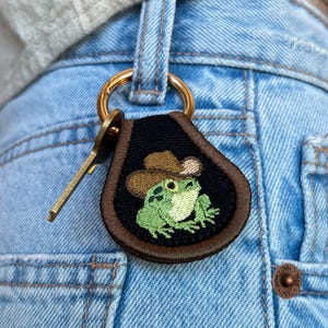 Accesorio/llavero con parche de rana vaquera con fondo oscuro, decoración para bolso o tirador.
