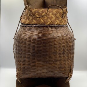 May include: A woven brown basket with a lid and a wooden base. The basket is made of natural materials and has a rustic look.