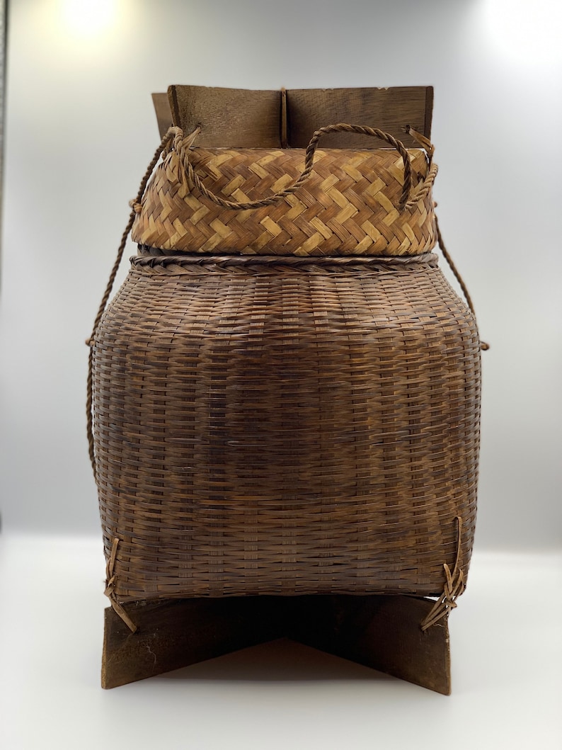 May include: A woven brown basket with a lid and a wooden base. The basket is made of natural materials and has a rustic look.
