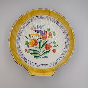 May include: A decorative, shell-shaped ceramic dish with a yellow scalloped rim. The center features a colorful bird and floral design with orange, green, and blue accents. A blue and white border encircles the central design.