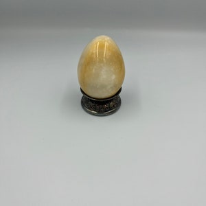 May include: A smooth, off-white alabaster egg sits on a decorative, silver-toned stand. The egg is slightly translucent and has a subtle sheen.