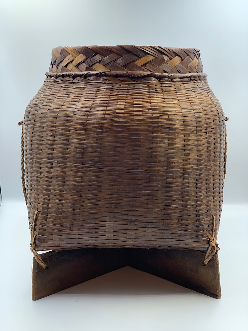 May include: A large, brown woven basket with a triangular wooden base. The basket is made of natural materials and has a woven pattern.