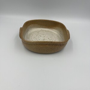 May include: A handmade, square ceramic baking dish with rounded corners. The dish has a speckled, off-white interior and a textured, brown exterior. It features small handles on either side, perfect for oven-to-table serving.