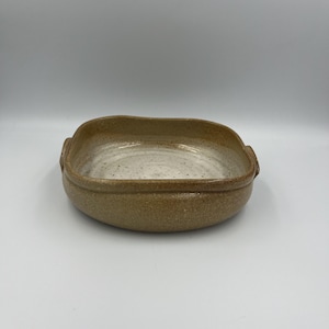 May include: A handmade, square ceramic baking dish with a light brown exterior and a speckled, off-white interior. The dish has rounded corners and small handles on each side. The overall aesthetic is rustic and earthy.