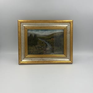 May include: A framed landscape painting featuring a river winding through a valley. The artwork has a gold and silver frame. The painting depicts trees, a small building, and a cloudy sky.