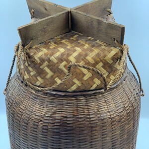 May include: A large, woven, brown basket with a lid made of woven straw and a wooden frame. The basket has two rope handles.
