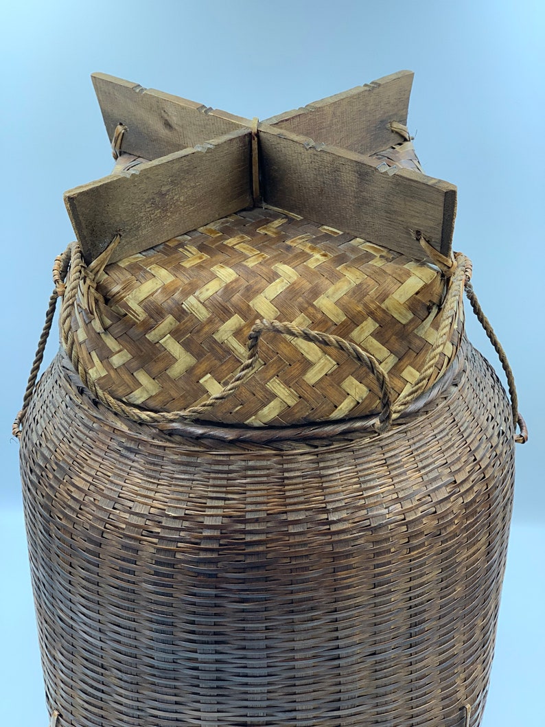 May include: A large, woven, brown basket with a lid made of woven straw and a wooden frame. The basket has two rope handles.