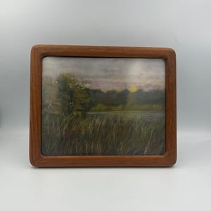 May include: A framed landscape painting depicting a marsh scene with tall grasses, a body of water, and a tree. The artwork features a sunset with a warm glow, and is encased in a wooden frame.