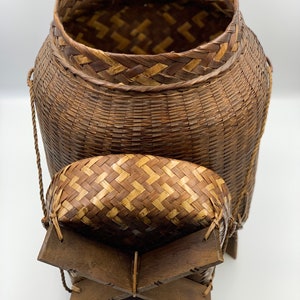 May include: A woven brown basket with a lid and a wooden base. The basket has a woven pattern and is made of natural materials.