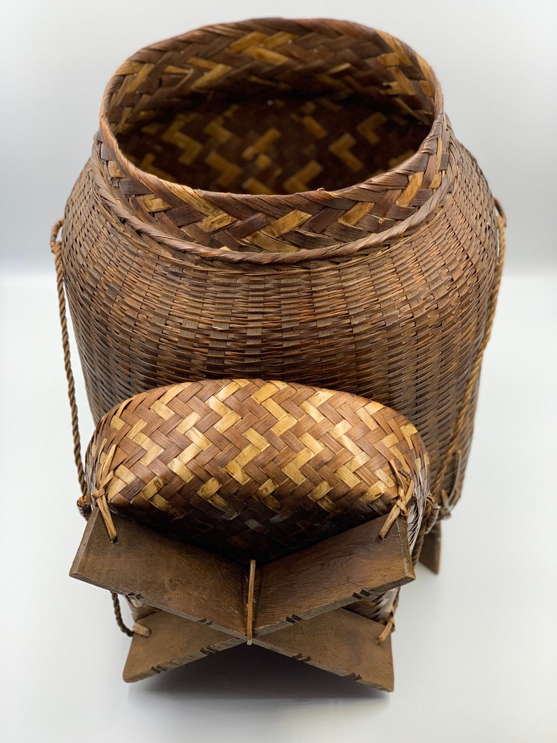 May include: A woven brown basket with a lid and a wooden base. The basket has a woven pattern and is made of natural materials.