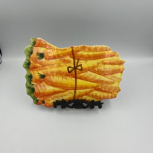 May include: A ceramic serving dish shaped like a bundle of carrots. The carrots are painted in shades of orange, yellow, and green. A brown ribbon is tied around the bundle.