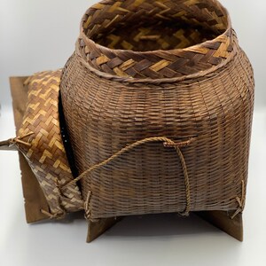 May include: A woven brown basket with a lid and a base made of woven wood. The basket has a rope handle and is sitting on a white surface.