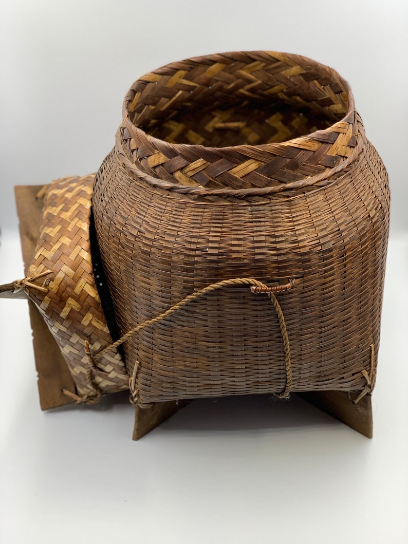 May include: A woven brown basket with a lid and a base made of woven wood. The basket has a rope handle and is sitting on a white surface.