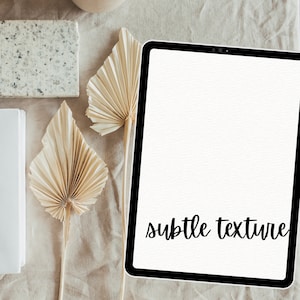 May include: A white tablet with a black border and a white background. The text "subtle texture" is written in black cursive on the screen. The tablet is on a beige surface with two dried palm leaves.