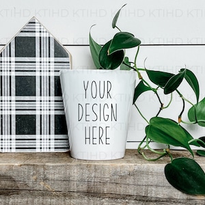 May include: White ceramic planter with the text "YOUR DESIGN HERE" on it. A green plant with trailing vines is in the planter. The planter is sitting on a wooden shelf with a black and white plaid house-shaped sign behind it.