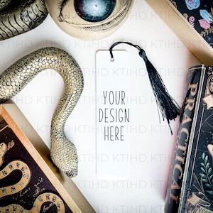Puede incluir: Un marcapáginas blanco con un borla negra y el texto "YOUR DESIGN HERE". El marcapáginas está sobre una superficie blanca con una serpiente dorada, un libro con una cubierta negra y una caja decorativa con un diseño floral.